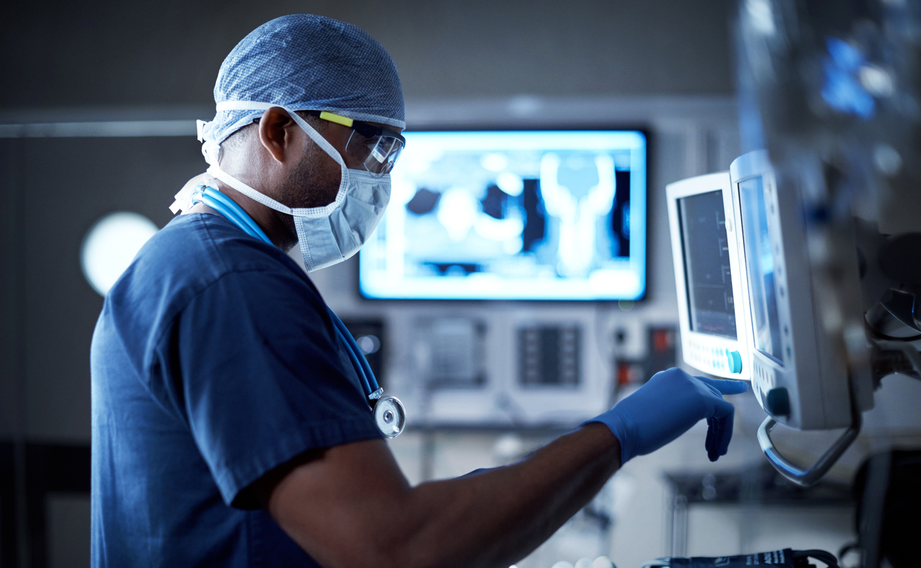 A woman wearing scrubs, a facemask, and latex gloves, uses a touch screen monitor