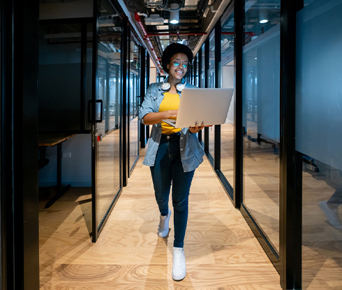 A woman walks down a row of servers with an open laptop in hand.