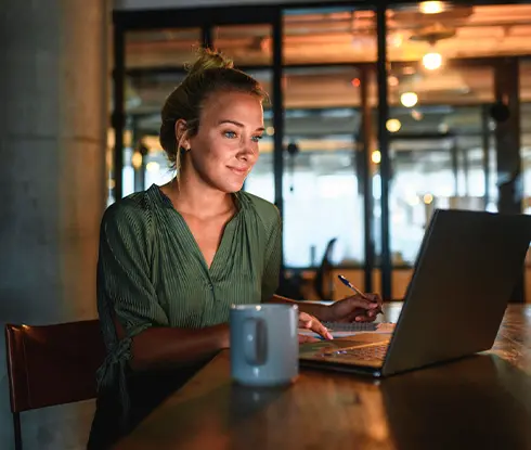 A woman with blonde hair sits at a desk working on a laptop.